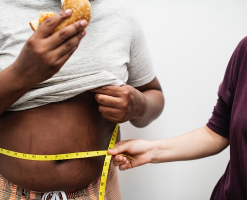 Woman holding a measuring tape around a fat man's waist while he is eating a burger