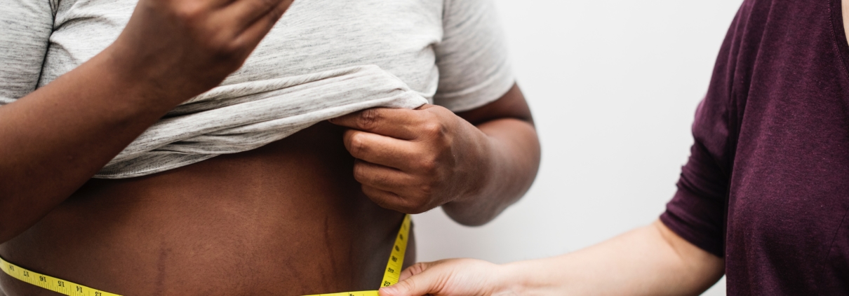 Woman holding a measuring tape around a fat man's waist while he is eating a burger