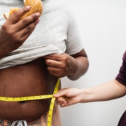 Woman holding a measuring tape around a fat man's waist while he is eating a burger