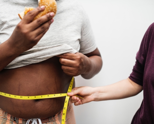 Frau hält ein Maßband um die Taille eines dicken Mannes, während er einen Burger isst