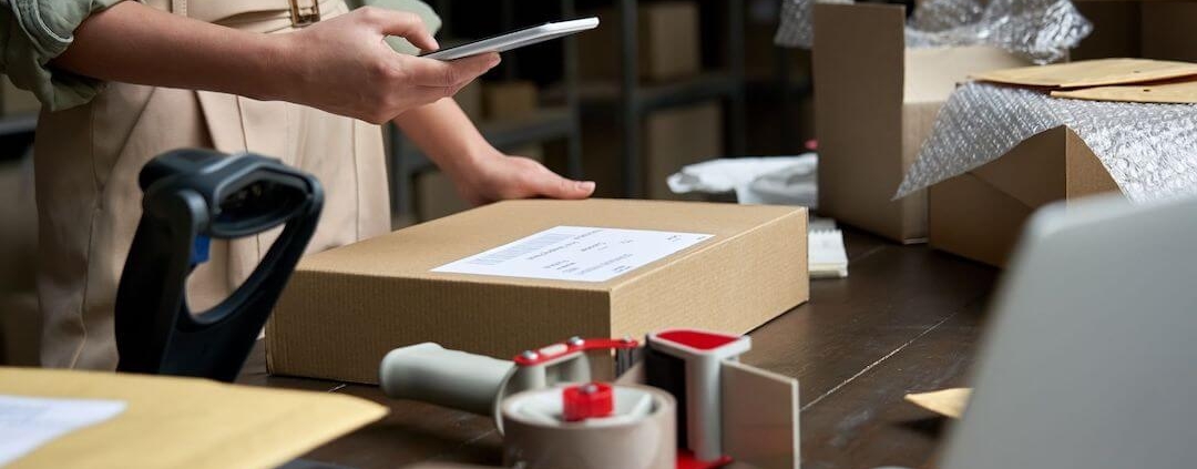 Frau hält ein Telefon über ein Paket auf einem Packtisch in einem Lagerhaus