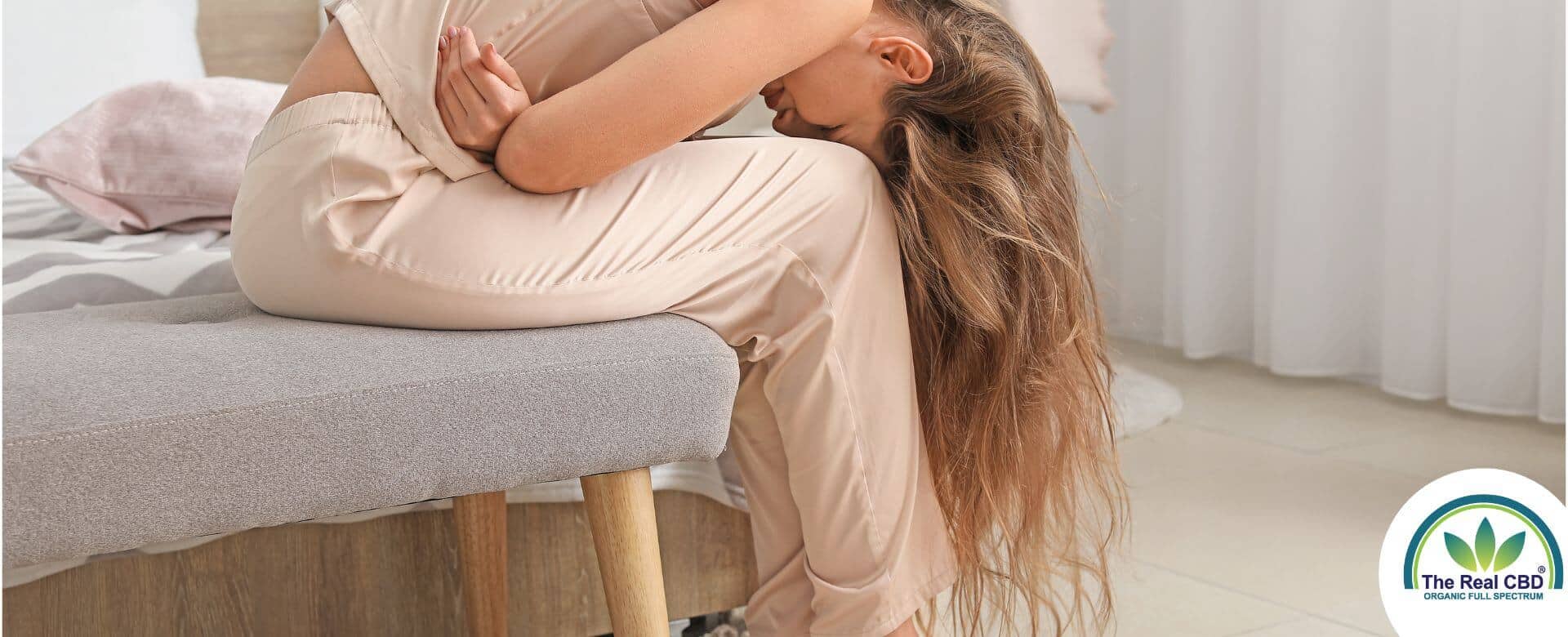 Femme aux cheveux longs assise sur un lit, pliée en deux par la douleur