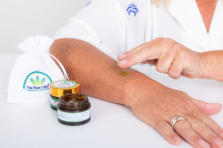 2 CBD balms, one without lid, next to a woman's arm. She is rubbing the balm on the skin - The Real CBD Brand